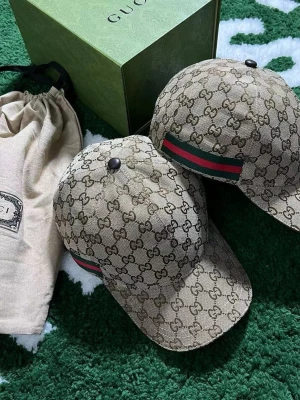 Beige keps från Gucci med GG-mönster - Säljer en beige keps från Gucci med det klassiska GG-monogrammönstret över hela kepsen. Kepsen har en böjd skärm och en grön-röd rand på sidan, typisk för Gucci. Materialet är canvasliknande tyg som känns lyxigt och trendigt.