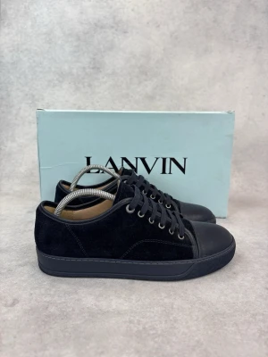 Svarta Lanvin sneakers i mocka och läder - Stilrena svarta sneakers från Lanvin med ovandel i mocka och tåparti i slätt läder. Skorna har svarta skosnören, metallöljetter och en svart gummisula. Perfekta för dig som gillar en clean och klassisk look.