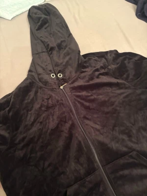 Svart velourhoodie från Gina Tricot - Gina Tricots mjuka och sköna svarta hoodie i velour är perfekt för en avslappnad stil.  Den har dragkedja framtill, stor huva med metallöljetter och fickor på magen. Det glansiga materialet känns lyxigt mot huden. Passar också för M-L