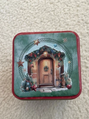 Julig plåtburk med dörrmotiv - Säljer en fyrkantig plåtburk i grönt och rött med ett mysigt julmotiv av en dekorerad dörr, gran och stjärnor. Perfekt för att förvara småsaker eller julgodis. Burken har detaljerade illustrationer och klassisk julkänsla.