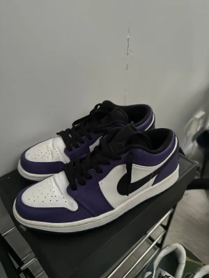 Nike Air Jordan 1 Low Court Purple - Snygga Nike Air Jordan 1 Low Court Purple sneakers med vit tåbox, lila och svarta detaljer samt svart swoosh. Skorna har klassisk låg siluett, svart snörning och ovandel i läder. Perfekta för dig som vill sticka ut med stilrena färgblock och ikonisk design.