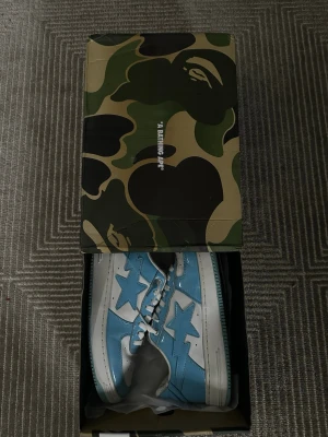 BAPE Bapesta blå och vita sneakers - Säljer ett par BAPE Bapesta sneakers i blått och vitt läder med den ikoniska stjärnloggan på sidan. Skorna har vita snören, perforerad tå och låg profil. Kommer i originalkartong med camomönster. Perfekta för dig som gillar streetwear och vill sticka ut.