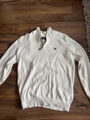 Vit stickad tröja från Denim Culture - Säljer en vit stickad tröja från Denim Culture med halv dragkedja och ståkrage. Tröjan har ett broderat mörkgrönt emblem på bröstet och ribbade muddar vid ärmslut och nederkant. Perfekt för dig som gillar klassisk och stilren look.