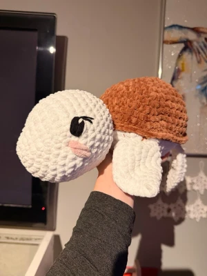 Virkad sköldpadda - Supergullig handvirkad sköldpadda i mjukt garn. Skölden är brun och kroppen vit, med svarta broderade ögon och rosa detalj på kinden. Perfekt som dekoration eller gosedjur. Gjord i tjockt, fluffigt material som känns mysigt att hålla i.