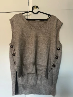 Beige stickad väst med knappar - Säljer en beige stickad väst med v-ringning och dekorativa knappar på sidorna. Västen har ribbade kanter och en längre, loose passform med slitsar i sidorna. Perfekt att styla över en skjorta eller t-shirt för en trendig lager-på-lager-look.