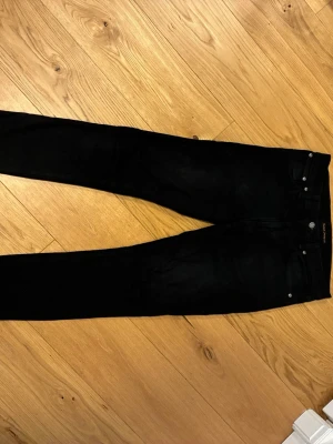 Svarta jeans från nudie - Säljer ett par svarta jeans från nudie. Jeansen har silverfärgade nitar och knapp, samt en diskret logga bak i midjan. Perfekta för en clean och stilren look.