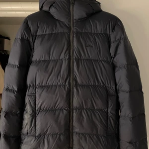 Vinterjacka från J.Lindeberg - Säljer en mörkblå pufferjacka från J.Lindeberg med huva och dragkedja framtill. Jackan har en stilren look med diskret logga på bröstet och två fickor på sidorna. Perfekt för kalla dagar när du vill hålla värmen och ändå se snygg ut.