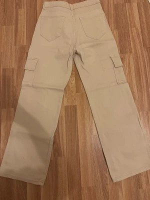 Beiga cargobyxor med raka ben - Säljer ett par beiga cargobyxor med raka ben och flera praktiska fickor på sidorna. Byxorna har två bakfickor och klassiska cargofickor med lock på båda benen. Perfekta för en avslappnad och trendig stil.