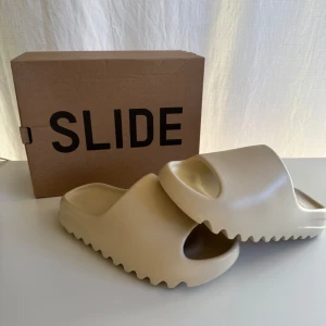 Adidas Yeezy Slide Bone - Själjer ett par snygga Yeezy Slides i  färgen Bone från Adidas. Perfekta för en bekväm dag på stranden eller hemma med stilren look. Storlek: 42 (US 8)Helt nya med lappen kvar. Priset är diskuterbart. Skriv vid minsta fundering!
