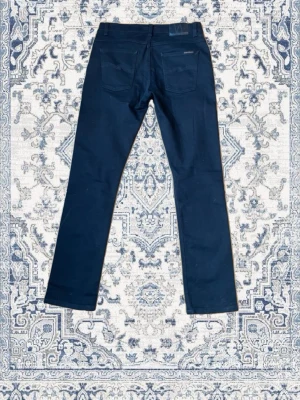 Svarta jeans från Nudie Jeans - Säljer ett par svarta jeans från Nudie Jeans med raka ben och klassiska bakfickor. Jeansen har diskret logga ovanför ena bakfickan och är tillverkade i ett slitstarkt denimtyg. Perfekta för dig som gillar stilrena och tidlösa plagg. Modell, Dude Dan.