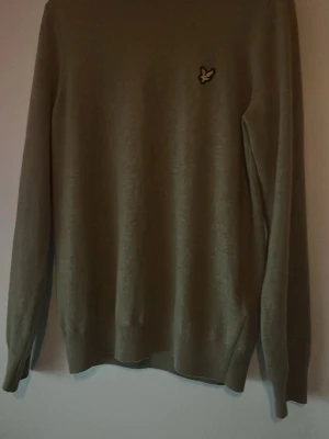 Beige stickad tröja från Lyle & Scott - Snygg beige stickad tröja från Lyle & Scott med deras klassiska gula fågel-logga på bröstet. Tröjan har rund halsringning, långa ärmar och ribbade muddar vid ärmslut och nederkant. Perfekt för lager-på-lager och enkel att matcha. Tröjan är köpt från Zalando men var för stor för mig. Den är därför bara använd en gång
