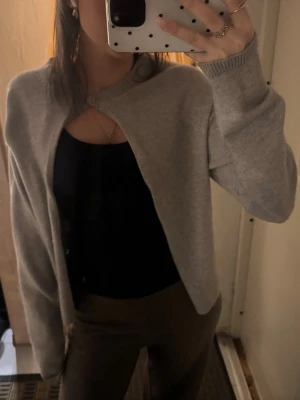 Ljusgrå cropped kofta med knapp - Snygg ljusgrå cropped kofta med en enkel knapp upptill. Koftan har lång ärm och är gjord i ett mjukt, tunt stickat material. Perfekt att slänga över ett linne eller topp för en chill look.