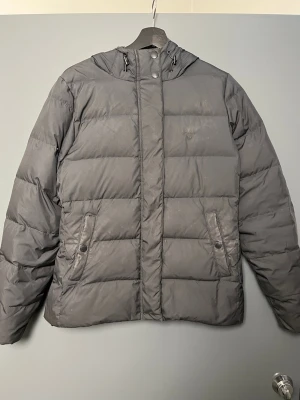 Grå dunjacka med huva från Gant - Snygg grå dunjacka från Gant med huva och dragsko. Jackan har två framfickor med knappstängning och stängs med både dragkedja och tryckknappar. Klassisk quiltad design som håller dig varm under kalla dagar. Perfekt för vintern.