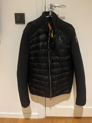 Svart cardiganjacka  från Parajumpers - Snygg svart cardiganjacka från Parajumpers med quiltad front och rygg i glansigt material samt stickade ärmar och sidor. Jackan har dragkedja framtill, bröstficka med zip och ikonisk gul detalj vid halsen. Perfekt för kyliga dagar. Negativt att de har lossnat från den man knäpper jackan med syns på bild och innerfickan så har kardborre grejen lossnar lite syns också även på bild annars är den i bra sick