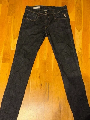 Replay anbass mörkblå jeans W29 - Snygga mörkblå jeans från Replay Hyperflex med klassisk femficksdesign och kontrastsömmar. Jeansen har en slim fit-modell och är tillverkade i stretchigt denim för extra komfort. Perfekta för dig som gillar stilrena och bekväma jeans. W29