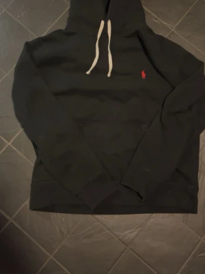 Svart hoodie från Polo Ralph Lauren - Klassisk svart hoodie från Polo Ralph Lauren med vit dragsko i huvan och den ikoniska röda loggan broderad på bröstet. Hoodien har ribbade muddar vid ärmslut och nederkant. Perfekt för en avslappnad och stilren look.