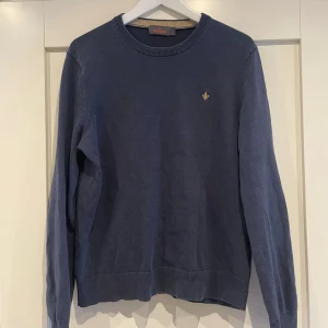 Mörkblå stickad tröja från Morris - Snygg mörkblå stickad tröja från Morris med rund halsringning och diskret guldfärgad logga broderad på bröstet. Tröjan har ribbade muddar vid ärmslut och nederkant. Perfekt för dig som gillar klassisk och stilren design.