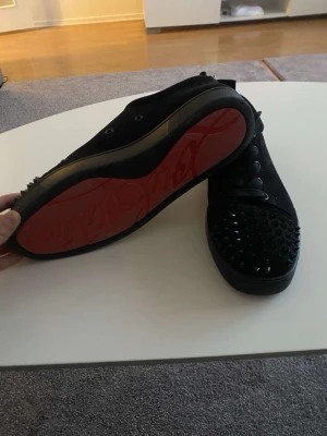 Svarta Louboutin sneakers med nitar, ÄKTA! - Säljer ett par svarta sneakers från Christian Louboutin med klassisk röd sula och svarta nitar på tån. Skorna är i mocka och har svarta skosnören. Perfekta för dig som vill sticka ut med en edgy och lyxig stil.