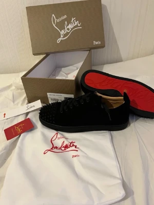 Svarta Louboutin sneakers med nitar - Säljer ett par svarta sneakers från Christian Louboutin med mattsvarta nitar på tån och klassisk röd sula. Skorna är låga och har ovandel i mocka med svarta skosnören. Kommer med originalkartong och dustbag.