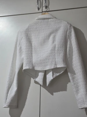 Vit kort kavaj i bouclétyg - Stilren vit kavaj i strukturerat bouclétyg med lång ärm och spetsig, asymmetrisk nederkant. Kavajen har en knapp framtill och klassisk krage. Perfekt för dig som vill ha en trendig och elegant look.