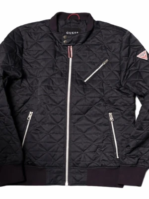 Svart quiltad bomberjacka Guess - Snygg svart quiltad bomberjacka från Guess med vita dragkedjor och ribbade muddar. Jackan har tre fickor med dragkedja och en liten logotyp på ärmen. Materialet är lätt glansigt och ger en modern look. Perfekt för dig som vill ha en stilren och trendig jacka.