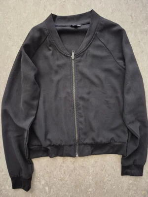 Bomberjacka - Stilren svart bomberjacka med hel dragkedja framtill och ribbade muddar vid ärmslut och midja. Jackan har en klassisk bombermodell med rundad halsringning och mjukt tyg. Perfekt för en enkel och clean look. Har en fläck.