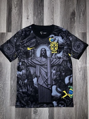 Nike Brasil svart grafisk fotbollströja - Unik svart fotbollströja från Nike med Brasil-logga och gult Nike märke. Framsidan har ett stort grafiskt tryck av Kristusstatyn och brasilianska motiv i grått, gult och grönt. Korta ärmar och rund hals. Perfekt för dig som vill sticka ut på planen.