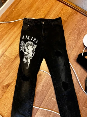 Svarta Amiri jeans med ängeltryck - Säljer ett par svarta jeans från Amiri med stort vitt ängeltryck och logga på vänster lår. slitna detaljer och hål på båda benen. Perfekta för dig som gillar streetwear och vill sticka ut med en unik design. OBS PRISET KAN DISKUTERAS 