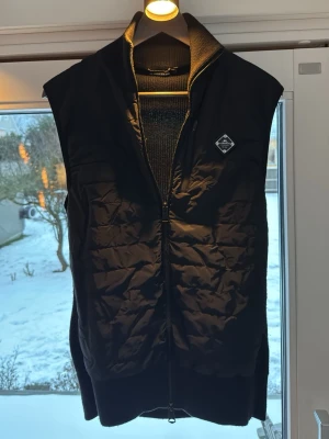 Svart dunväst från J.LINDEBERG - Snygg svart dunväst från J.Lindeberg, quiltad framsida och ribbade sidor. Västen har dragkedja framtill och en liten logotyp på bröstet. Perfekt att slänga över en hoodie eller tröja för extra värme. Klassisk och stilren design.