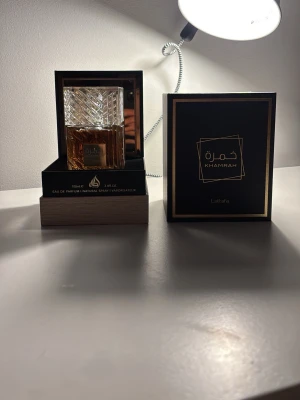 Lattafa Khamrah Eau de Parfum 100ml - Lattafa Khamrah Eau de Parfum kommer i en lyxig, fyrkantig glasflaska med ett elegant mönster och gulddetaljer. Doften har en varm bärnstensfärg och flaskan levereras i en svart och guld ask som ger en exklusiv känsla. Perfekt för dig som gillar unika och sofistikerade parfymer. 100/100ml