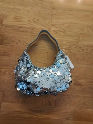 Silvrig väska med paljetter - Glittrig handväska i silver paljetter. Helt oanvänd med lapp kvar. Säljer för att jag har en liknande. Annars super fin! 
