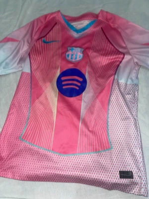 Barça rosa matchtröja Nike - Snygg rosa FC Barcelona matchtröja från Nike med blå detaljer och vita inslag. Tröjan har klubbmärke, Nike-logga och Spotify-logga på bröstet. Tillverkad i lätt och ventilerande funktionsmaterial, perfekt för fotboll eller träning.