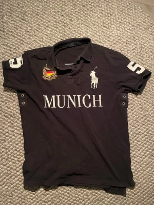 Svart polo t-shirt Ralph Lauren Munich - Klassisk svart t-shirt från Ralph Lauren med stor vit 'MUNICH'-text och polospelare på bröstet. Nummer 5 på både ärm och rygg, samt broderad patch med tysk flagga. Skön bomull och kortärmad modell med krage och knappar.