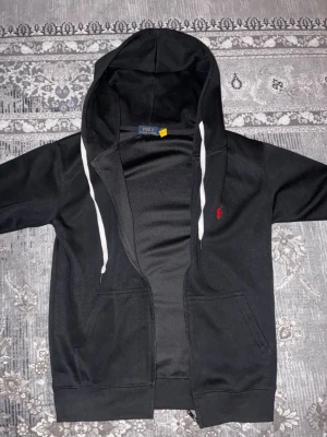 Svart hoodie från Polo Ralph Lauren - Säljer en svart hoodie från Polo Ralph Lauren med dragkedja, vita snören i huvan och den klassiska röda loggan broderad på bröstet. Hoodien har två fickor framtill och är gjord i mjukt bomullsmaterial. Perfekt för en avslappnad och stilren look.