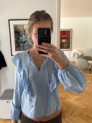 Ljusblå blus med volanger - Snygg ljusblå blus med långa ärmar och volangdetaljer längs axlar och bröst. Blusen har v-ringning med knytband.