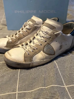 Philippe Model vita sneakers med mockadetaljer - Snygga vita sneakers från Philippe Model med beige mockadetaljer och logga på sidan. Skorna har vita snören med tryckt text och en klassisk låg siluett. Ovandelen är i läder och mocka, med svart detalj vid hälen. Perfekta för en trendig streetstyle-look