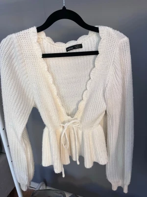 Vit stickad omlottopp från SHEIN - Söt vit stickad omlottopp från SHEIN med volangkant och knytband i midjan. Tröjan har lång ärm, ribbstickad struktur och peplumdetalj nedtill. Perfekt för dig som gillar romantisk och trendig stil. En av trådarna har åkt av så jag har satt en säkerhetsnål. De går att sy fast enkelt 