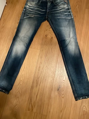 Blå slitna jeans från Replay - Säljer ett par blå jeans från Replay modell Anbass med snygga slitningar och riktigt feta tvättade detaljer. Jeansen är i materialet kraftig denim i bomull. Perfekta för en avslappnad och trendig look. Säljer då de är för små tyvärr. Storlek W33 L34
