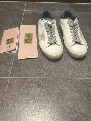 Golden Goose vita sneakers med stjärna - Golden Goose sneakers i vitt läder med grå mockastjärna på sidan, grå skosnören och blå detalj vid hälen. Klassisk låg modell med GGDB/STAR-tryck i blått på sidan. Snygg vintagekänsla och robust sula.