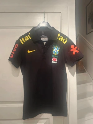 Svart Brasil Nike pikétröja - Säljer en svart pikétröja från Nike med Brasilien fotbollslandslagets logga på bröstet. Tröjan har gula och röda sponsorloggor på ärmar och axlar, samt klassisk krage och knappar framtill. Perfekt för dig som gillar fotboll och vill ha en sportig look.