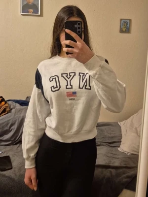Vit sweatshirt med NYC-tryck - Vit sweatshirt med svart NYC-tryck och liten USA-flagga framtill. Tröjan har svarta detaljer på axlarna och är i en klassisk, avslappnad passform med ribbade muddar vid ärmslut och midja. Perfekt för en chill och sportig look.