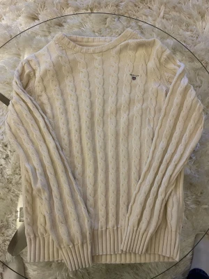 Kabelstickad offwhite tröja från Gant - Kabelstickad tröja i offwhite från Gant med rund halsringning och ribbade muddar. Tröjan har ett diskret Gant-logo broderat på bröstet och är gjord i mjukt material, perfekt för lager på lager. Klassisk och tidlös design som passar till allt.