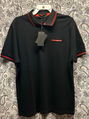 Svart polotopp från Prada  - Svart polotopp från Prada med röda ränder på krage och ärmslut samt en röd logga på bröstet. Toppen har korta ärmar och en half zip-dragkedja framtill. Snygg och stilren design som passar till mycket.