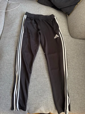 Svarta Adidas träningsbyxor - Säljer ett par svarta träningsbyxor från Adidas med klassiska vita ränder längs benen och Adidas-logga på låret. Byxorna har resår i midjan och är tillverkade i ett mjukt, stretchigt material som passar perfekt till träning eller chill.