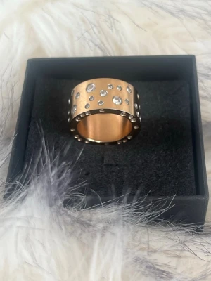 Bred guldfärgad ring med stenar - Snygg  ring från Edblad i guldfärgad metall med flera klara stenar i olika storlekar runt hela ringen. Ringen har en modern och chunky design som verkligen sticker ut. Perfekt för dig som gillar statement-smycken och vill ha något som syns.