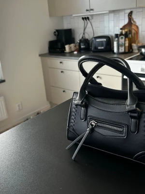 Svart handväska med dragkedja - Snygg svart handväska i skinnimitation med dubbla handtag och dekorativ dragkedja framtill. Väskan har en stilren design och passar perfekt för dig som gillar minimalistiska accessoarer. Perfekt storlek för det viktigaste och enkel att matcha med olika outfits.