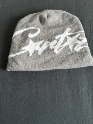 Cortez mössa - Säljer en grå mössa med vit graffiti-inspirerad text och stjärndetaljer framtill. Mössan är mjuk och stretchig, perfekt för dig som gillar streetwear och vill ha en cool accessoar till hösten eller vintern.