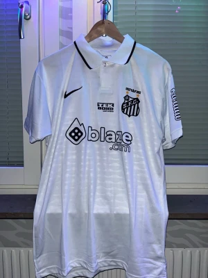 Vit Santos FC matchtröja Nike storlek S  - Snygg vit matchtröja från Santos FC med svart Nike-logga och klubbmärke broderat på bröstet. Tröjan har klassisk krage, tryckta sponsorer och detaljer i svart. Perfekt för dig som älskar fotboll och vill ha en stilren supportertröja.