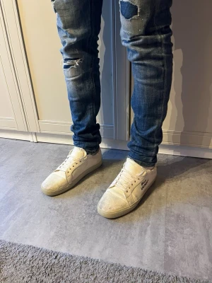 Vita Saint Laurent sneakers - Säljer ett par klassiska vita Saint Laurent sneakers med lågt skaft och snörning. Skorna har en stilren design med Saint Laurent-loggan på sidan och är tillverkade i canvas med gummisula. Perfekta för en clean och trendig look.