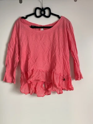 Rosa volangblus med knappar bak - Söt rosa blus med volangkant nertill och knappar längs ryggen. Blusen har rund halsringning och trekvartsärm. Tillverkad i ett lätt och luftigt tyg, perfekt för en avslappnad och trendig look. Skrynklig på bild & går att stryka fint! 